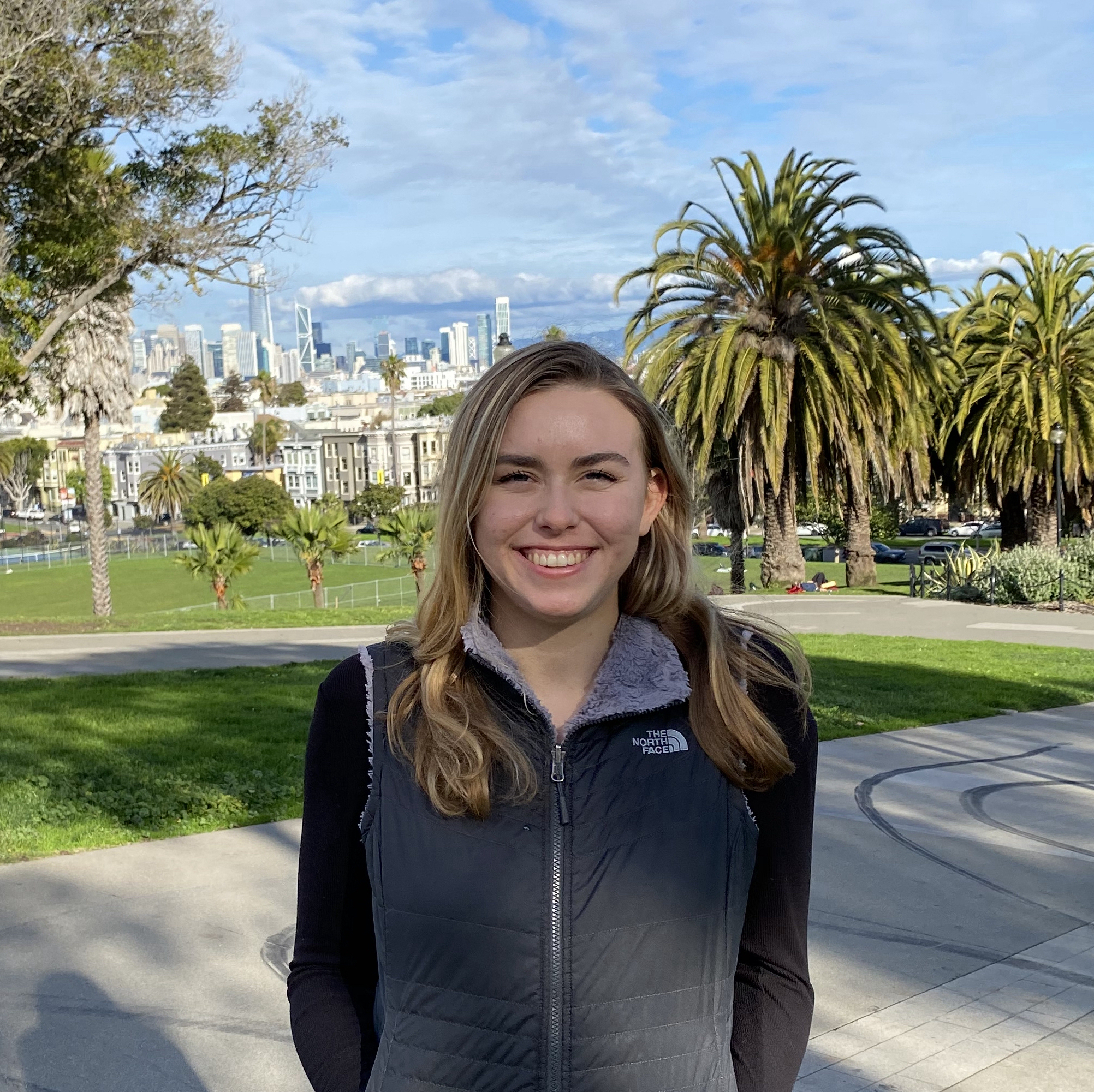Claire is standing in front of a park with palm trees. In the background you can see the San Francisco skyline. She is wearing a black shirt and a vest.