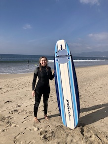 Claire posing with a surfboard with blue stripes in front of the ocean.
